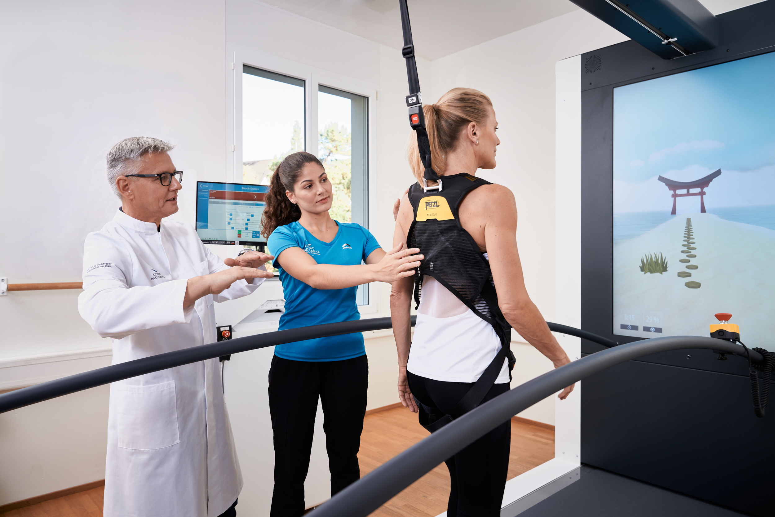Eine Frau in Sportkleidung benutzt ein Rehabilitations-Laufband mit Gurtzeug. Ein Arzt und ein Therapeut assistieren ihr. Auf dem Bildschirm wird ein ruhiger virtueller Weg angezeigt.