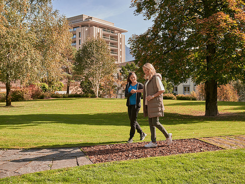 Zwei Frauen spazieren und unterhalten sich in einem sonnigen Park, umgeben von grünem Gras und hohen Bäumen. Ein modernes Gebäude im Hintergrund verleiht dem Bild einen urbanen Touch.