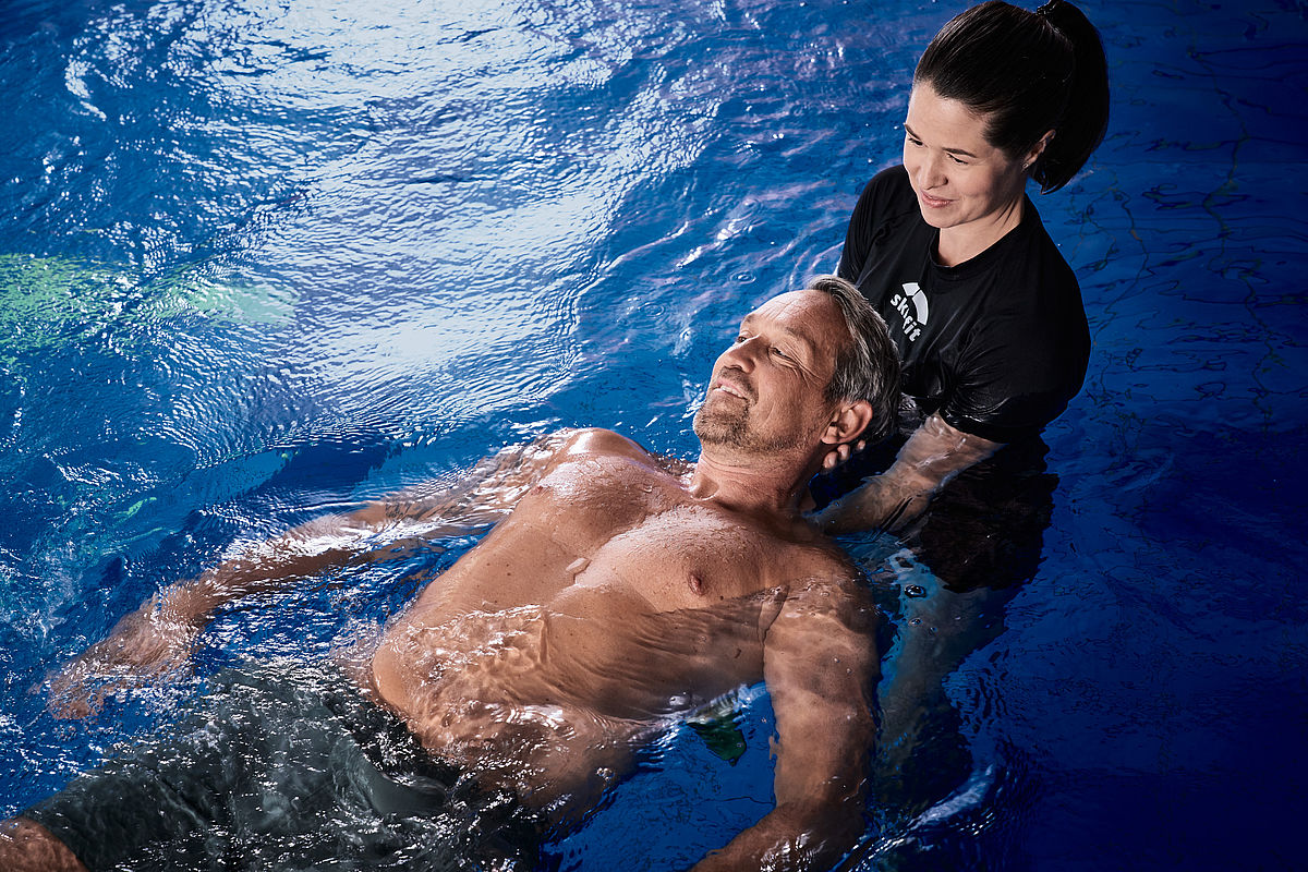 Ein Mann schwimmt friedlich auf dem Rücken in einem Pool, gestützt von einer lächelnden Frau. Die Szene vermittelt Entspannung und Vertrauen in einer beruhigenden blauen Wasserumgebung.