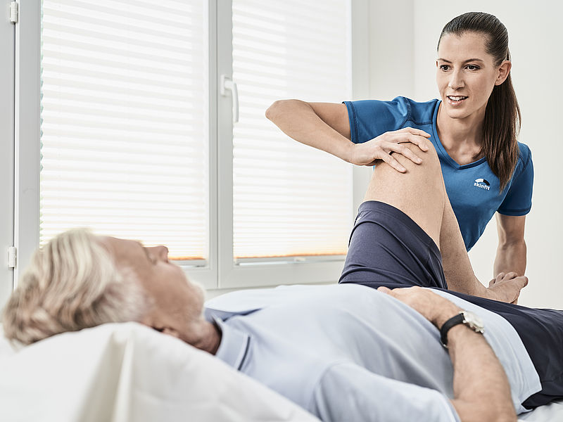 Eine Physiotherapeutin hilft einem älteren Mann in einer Klinik, indem sie sein Bein sanft dehnt. Die Umgebung ist hell und klinisch und vermittelt Sorgfalt und Professionalität.