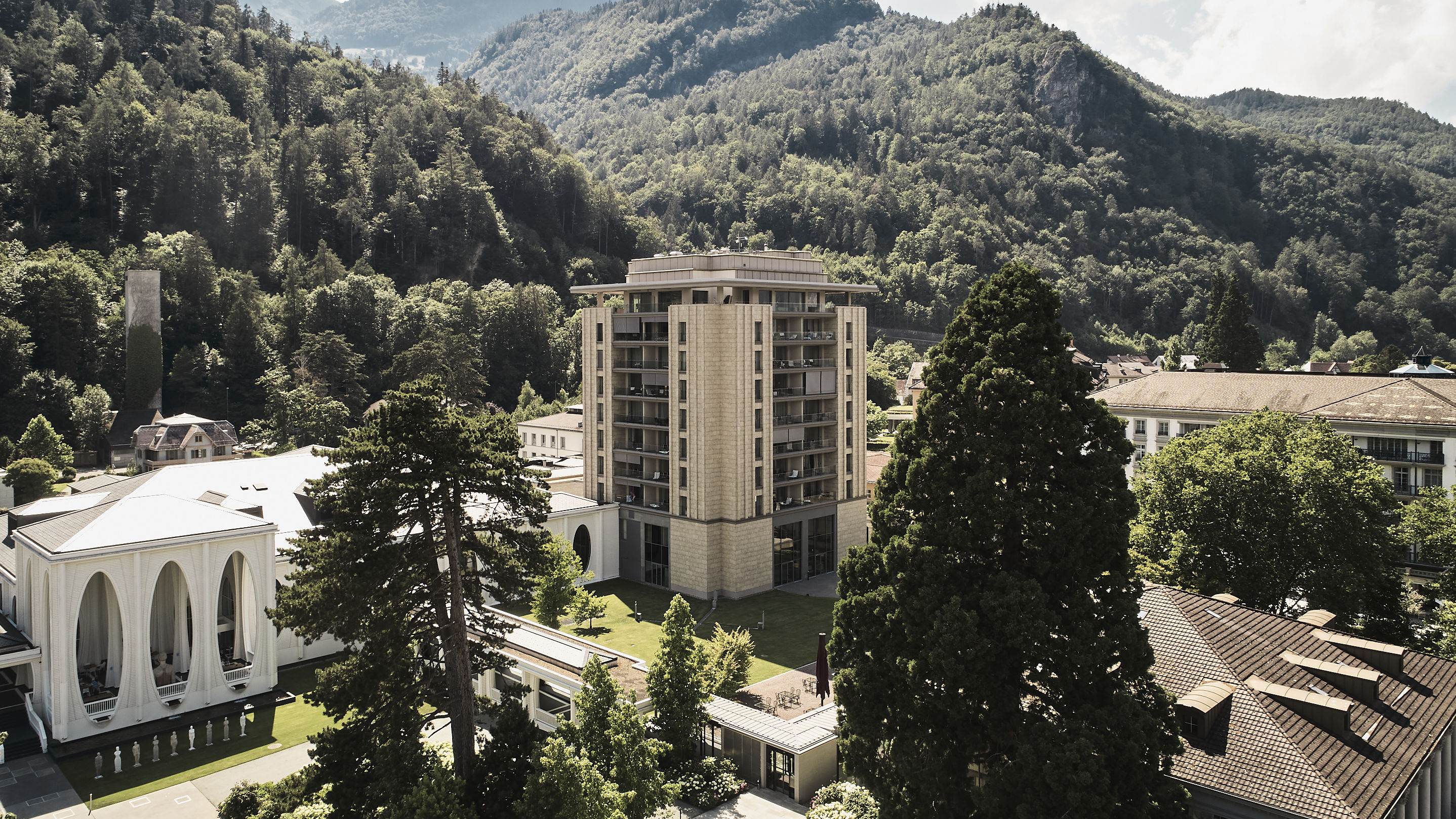 Ein hohes, beigefarbenes Gebäude steht inmitten üppig grüner Bäume, vor einer Bergkulisse und unter einem teilweise bewölkten Himmel. In der Nähe sorgt ein weißes Gebäude für Eleganz.