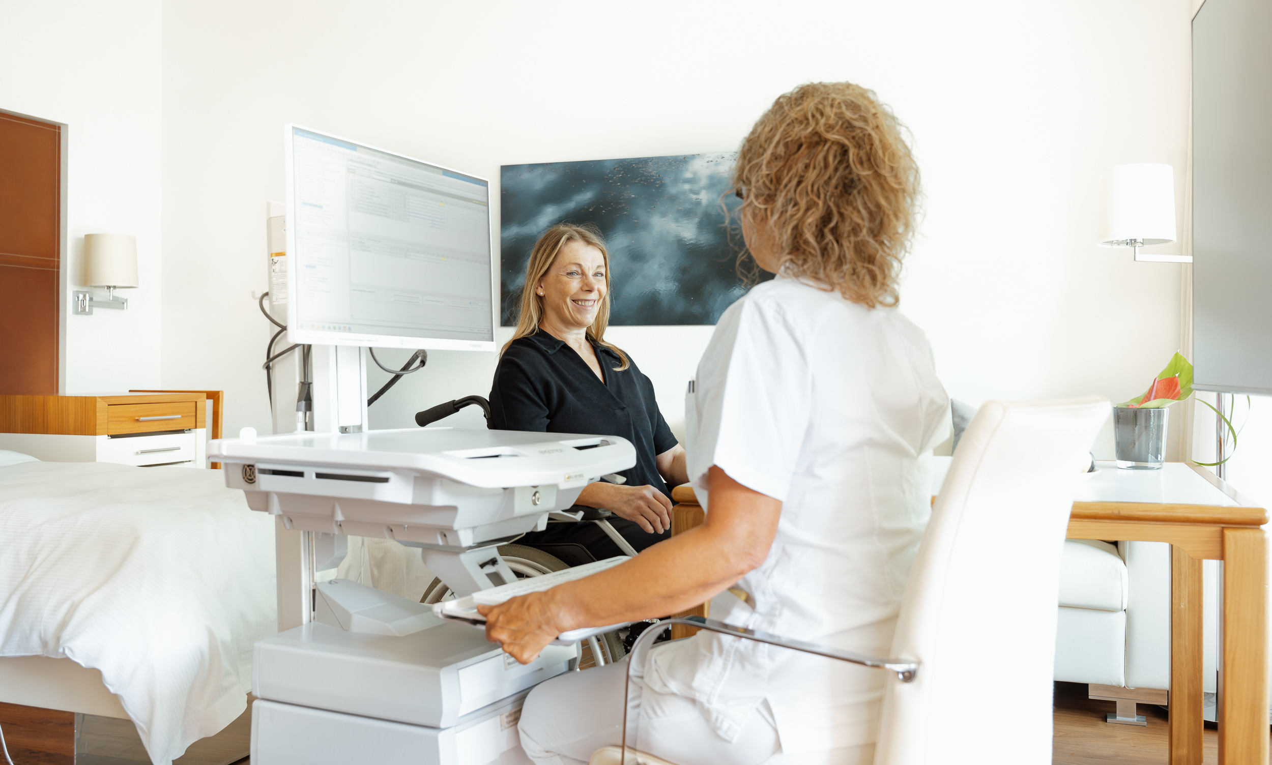 Eine Krankenschwester in weißer Uniform sitzt mit einer älteren Frau auf einem Liegestuhl und schaut auf ein Klemmbrett. Die Frau lächelt warmherzig. Auf einem Tisch in der Nähe stehen Frühstücksartikel.