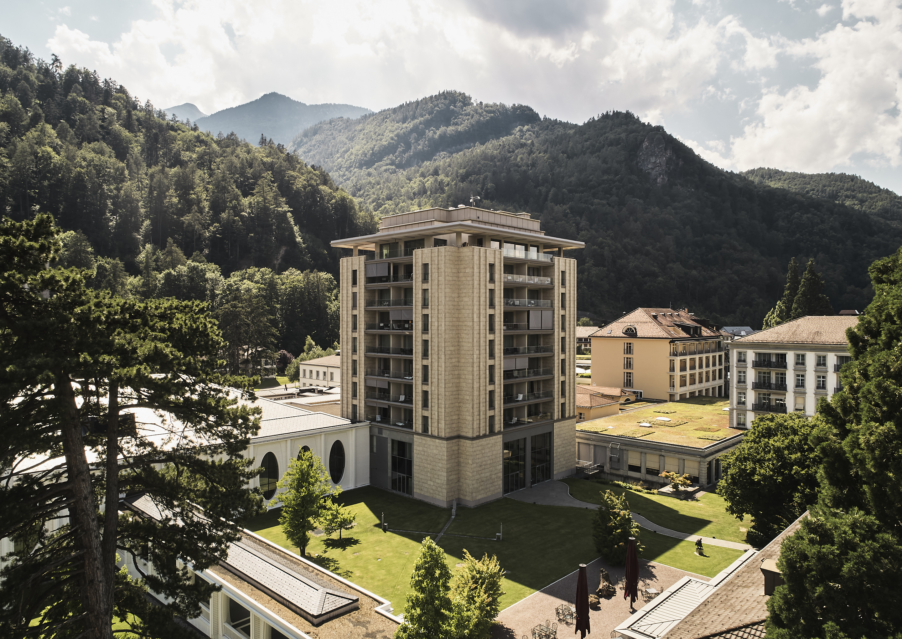 Ein hohes, modernes Gebäude mit Balkonen steht inmitten von üppigem Grün und Bergen unter einem wolkenverhangenen Himmel und vermittelt eine ruhige und malerische Atmosphäre.